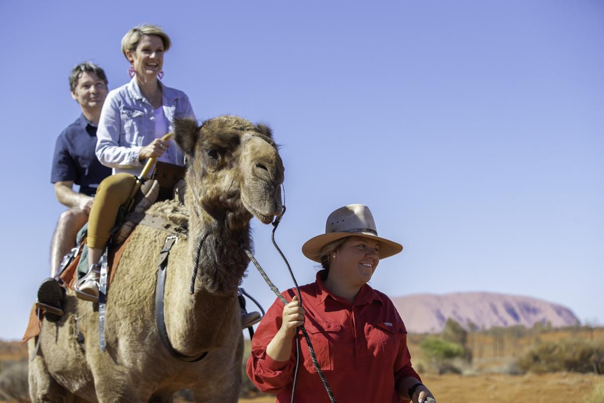 image of two people on camel being led by a guide with Uluru in background