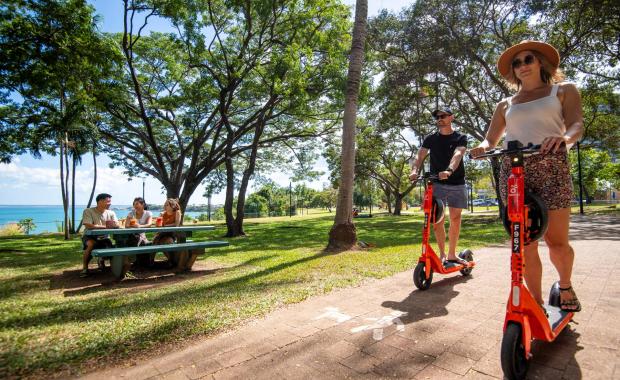 Man and woman on scooters riding along Darwin Esplanade. People enjoying picnic in the background.